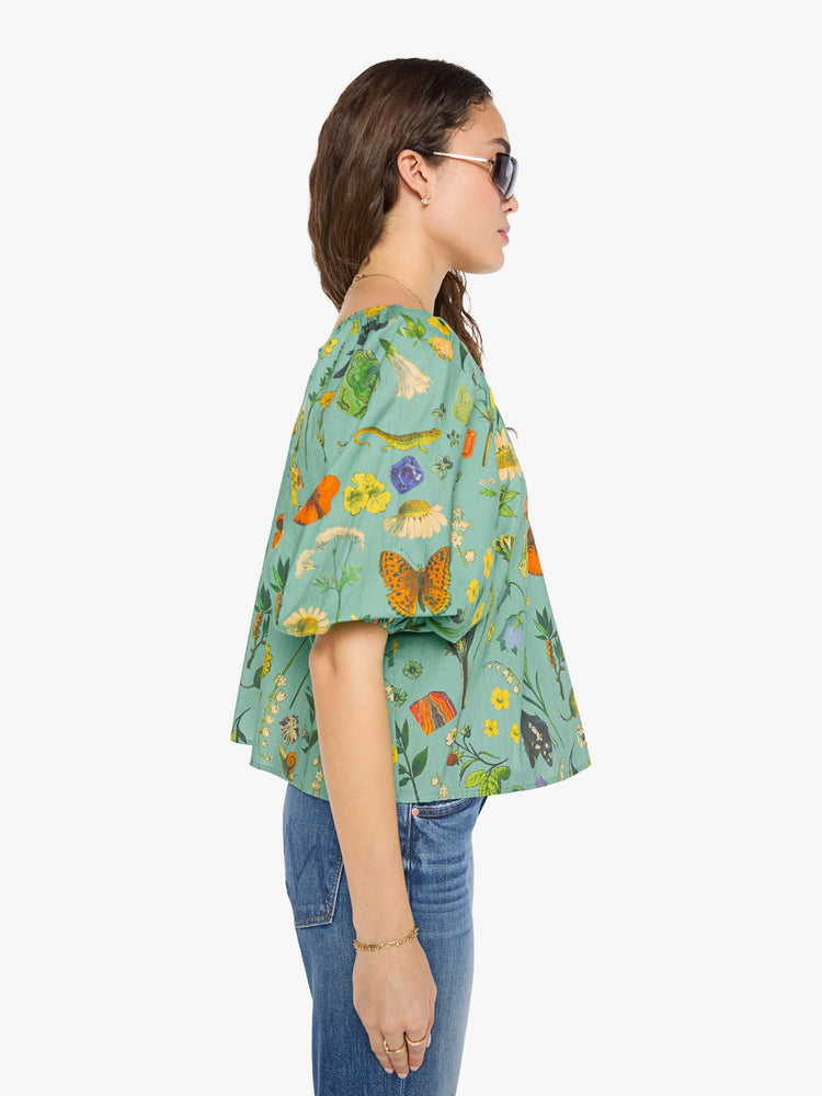 Side view of a woman in a teal-green with oversized printed flowers, gems, and creatures throughout flowy blouse.