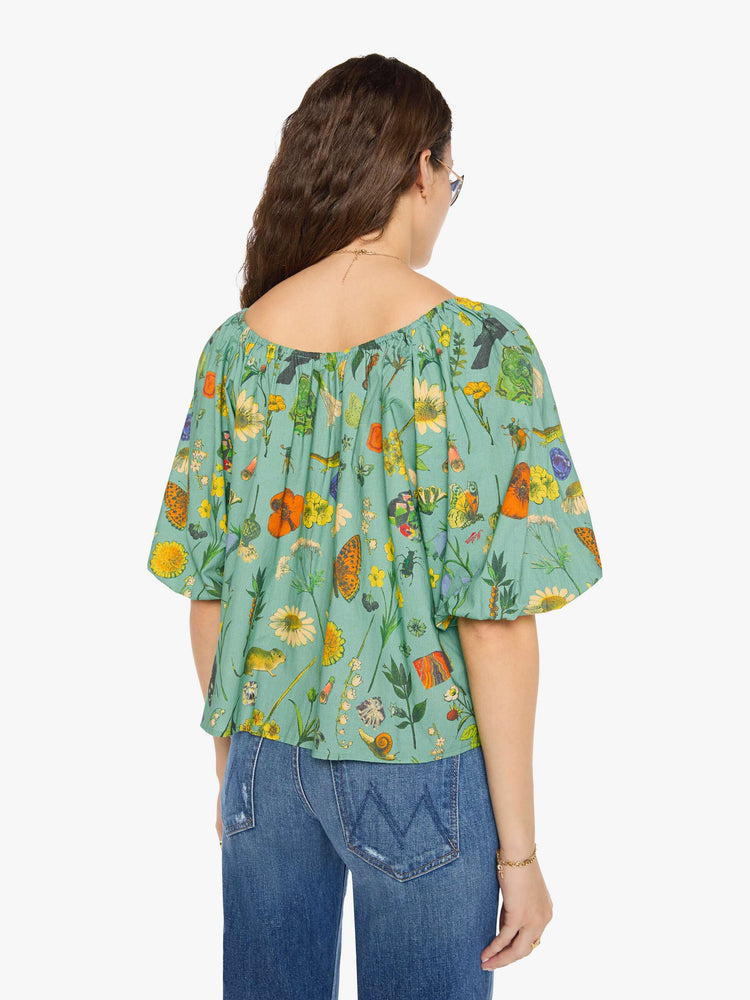 Back view of a woman in a teal-green with oversized printed flowers, gems, and creatures throughout flowy blouse.