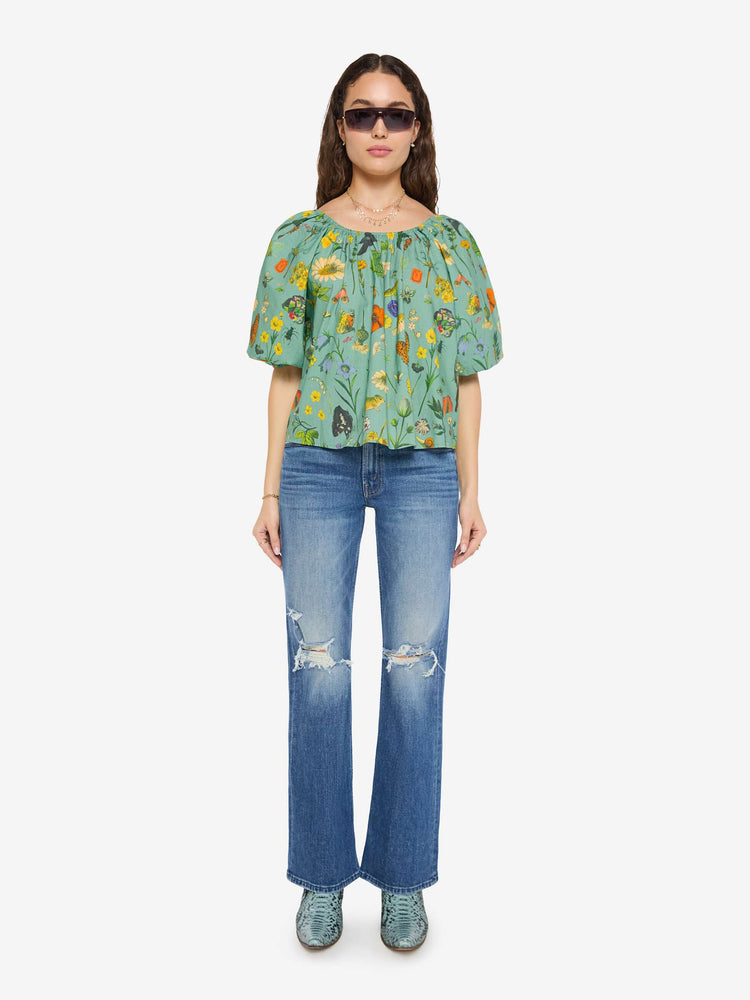 Front full body view of a woman in a teal-green with oversized printed flowers, gems, and creatures throughout flowy blouse.