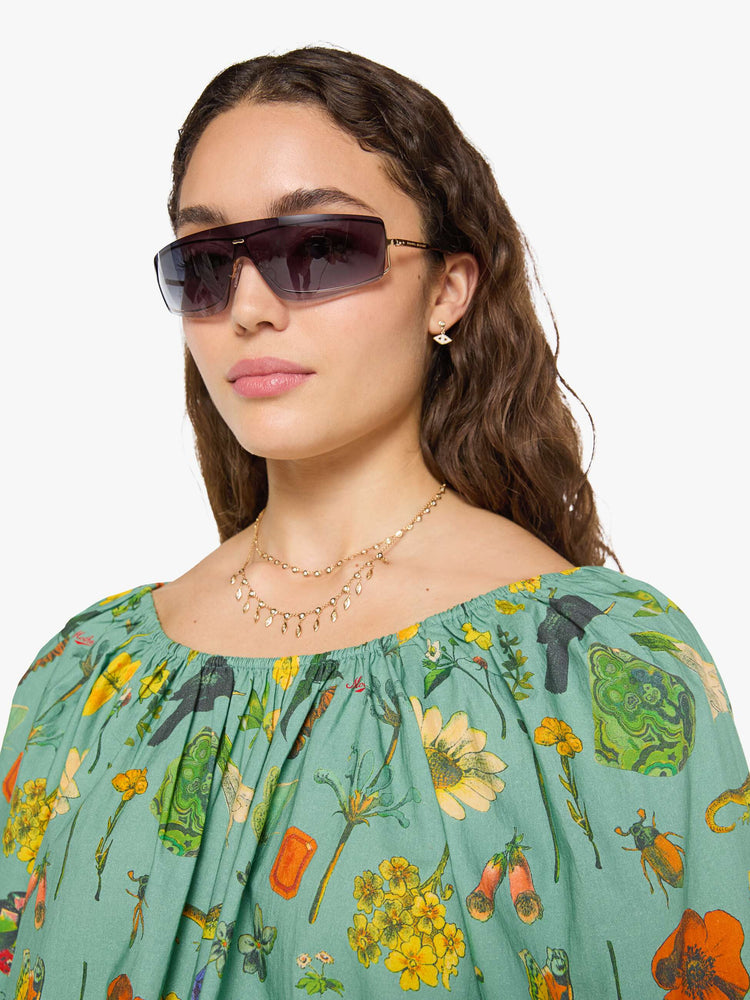 Close up view of a woman in a teal-green with oversized printed flowers, gems, and creatures throughout flowy blouse.