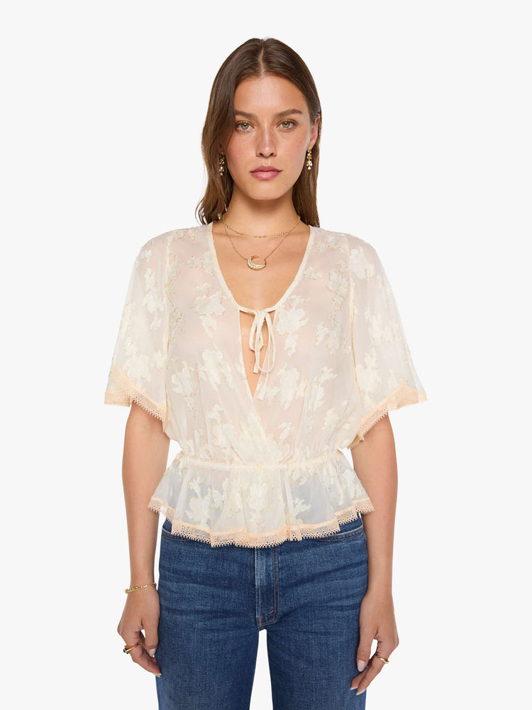 Front view of woman in a peach lace sheer blouse with a deep V-neck that ties, elbow-length flutter sleeves, an elastic waist and a flowy hem.