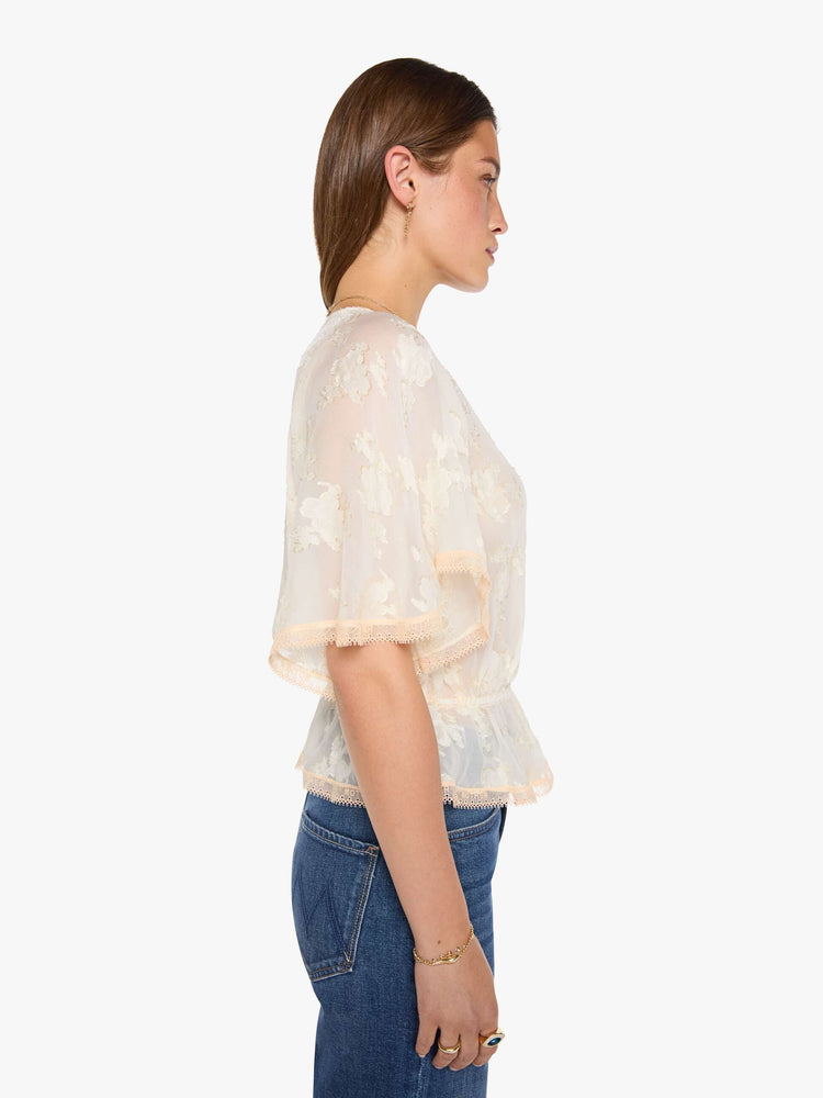 Side view of woman in a peach lace sheer blouse with a deep V-neck that ties, elbow-length flutter sleeves, an elastic waist and a flowy hem.