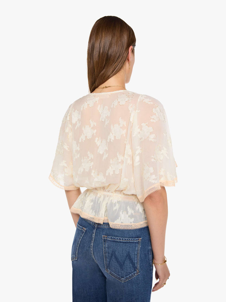 Back view of woman in a peach lace sheer blouse with a deep V-neck that ties, elbow-length flutter sleeves, an elastic waist and a flowy hem.