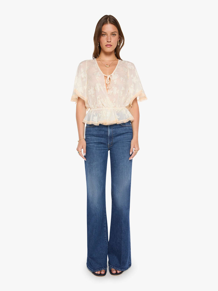 Full body view of woman in a peach lace sheer blouse with a deep V-neck that ties, elbow-length flutter sleeves, an elastic waist and a flowy hem.