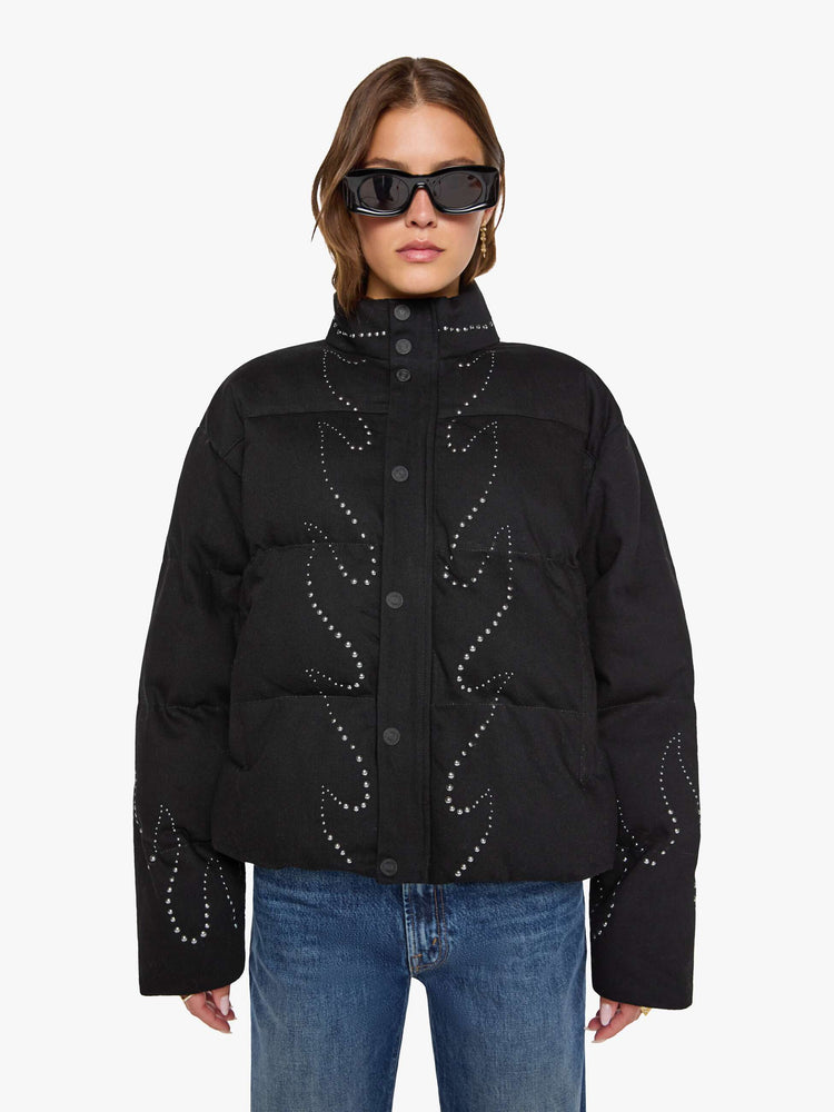 Front view of a woman in a black with flame studded down th middle  boxy puffer jacket with a stacked collar, drop shoulders, slit pockets, a zip closure that buttons and a slightly cropped fit. 