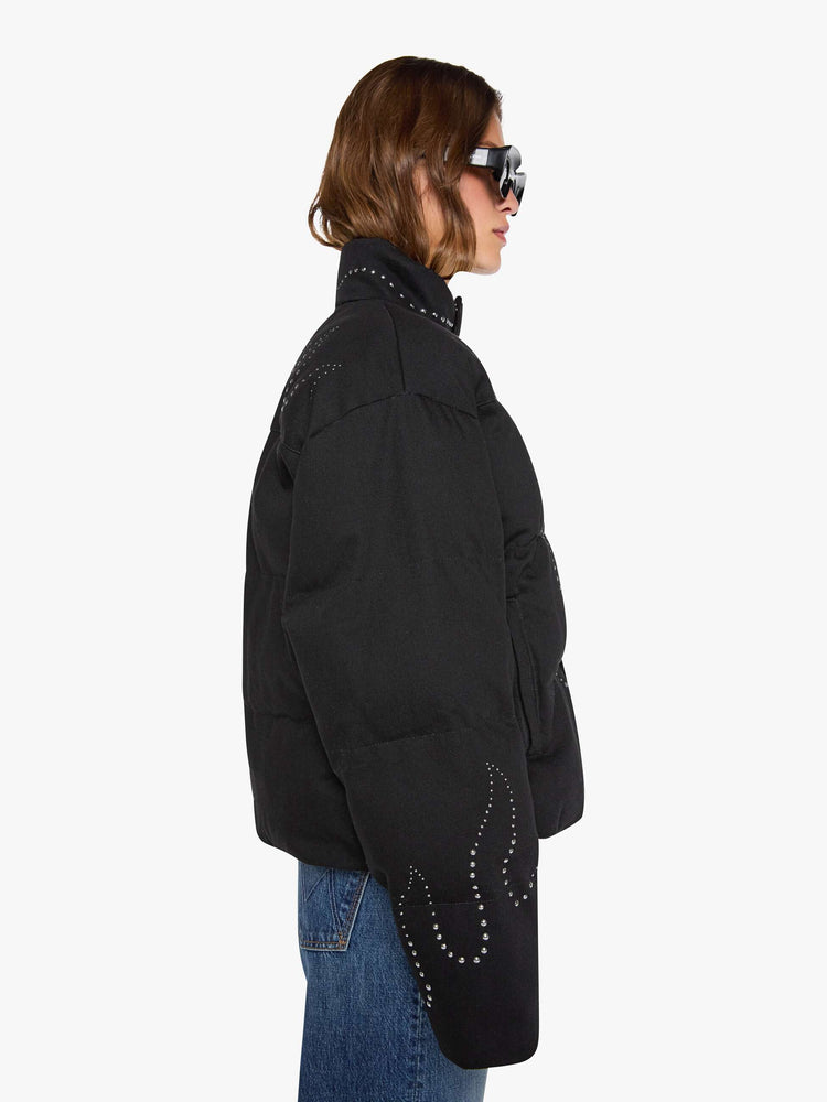 Side view of a woman in a black with flame studded down th middle  boxy puffer jacket with a stacked collar, drop shoulders, slit pockets, a zip closure that buttons and a slightly cropped fit. 