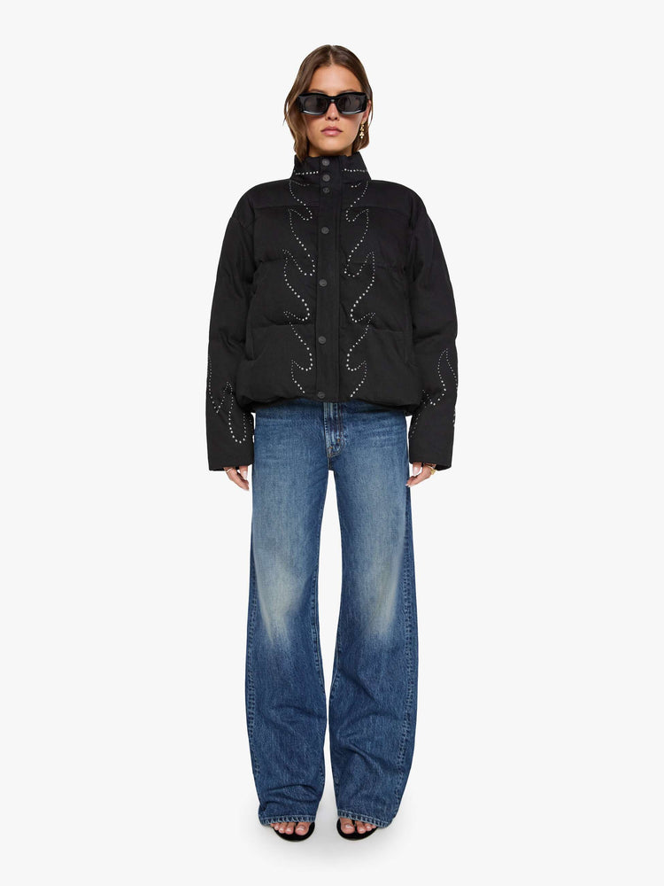 Front full body view of a woman in a black with flame studded down th middle  boxy puffer jacket with a stacked collar, drop shoulders, slit pockets, a zip closure that buttons and a slightly cropped fit. 