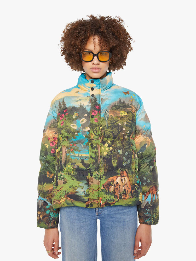 Front view of a woman in a puffer jacket with a high collar, zip and snap closure, slit pockets in a quilted fabric with a colorful pastoral graphic.