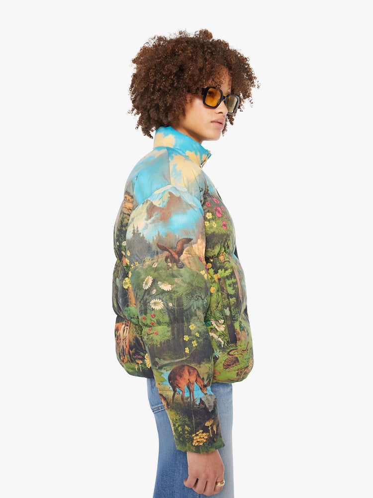 Side view of a woman in a puffer jacket with a high collar, zip and snap closure, slit pockets in a quilted fabric with a colorful pastoral graphic.