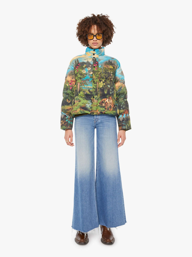 Full body view of a woman in a puffer jacket with a high collar, zip and snap closure, slit pockets in a quilted fabric with a colorful pastoral graphic.