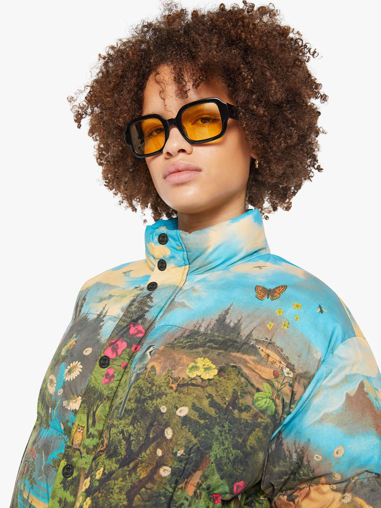 Close up view of a woman in a puffer jacket with a high collar, zip and snap closure, slit pockets in a quilted fabric with a colorful pastoral graphic.