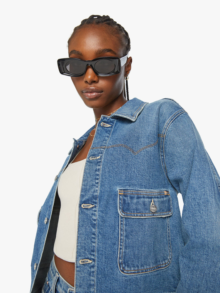Close up view of a woman boxy denim jacket with Western-inspired stitching across the chest, drop shoulders and oversized patch pockets in a med blue wash.