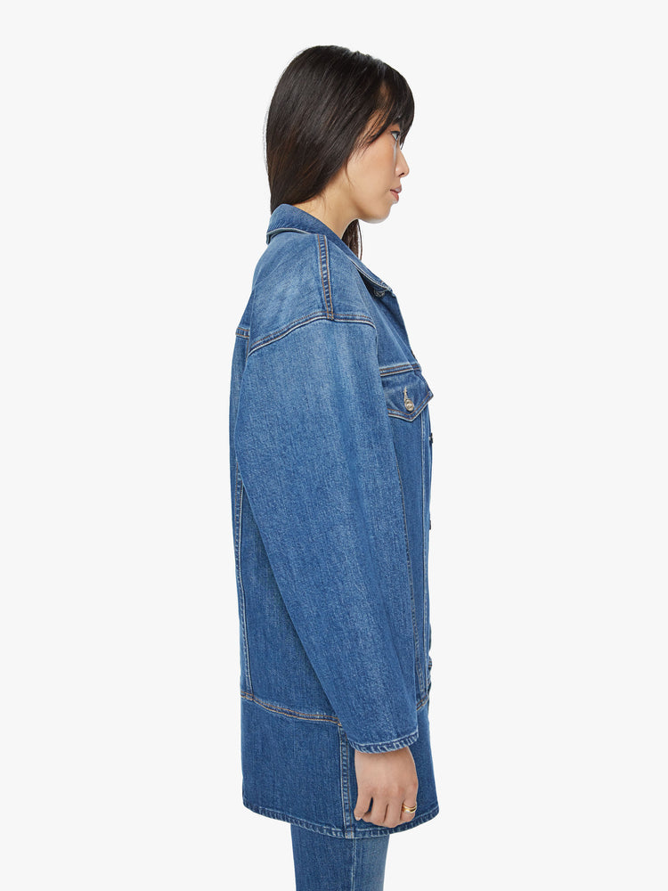 Side view of a woman elongated denim jacket with drop shoulders, boxy wing sleeves and a curved, thigh-length hem in classic blue wash.