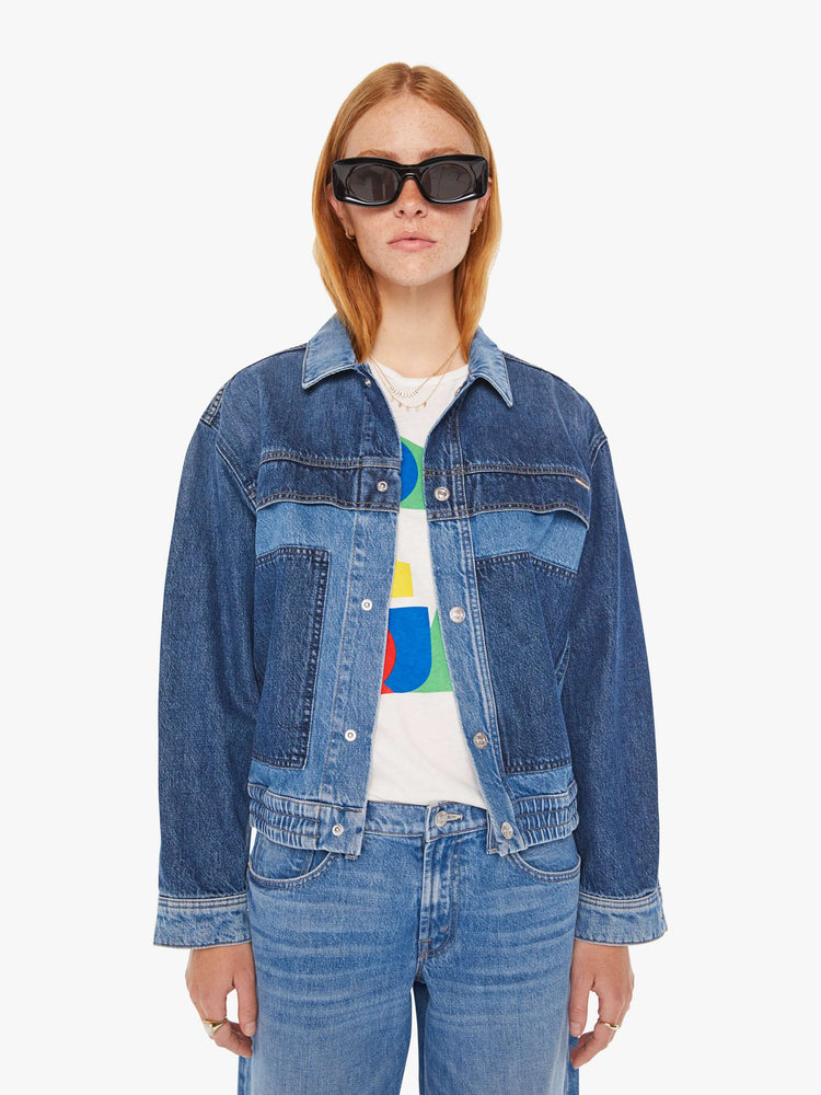 Front view of a woman mid blue denim jacket with drop shoulders, patch pockets and an elastic hem at the waist with darker details on chest.