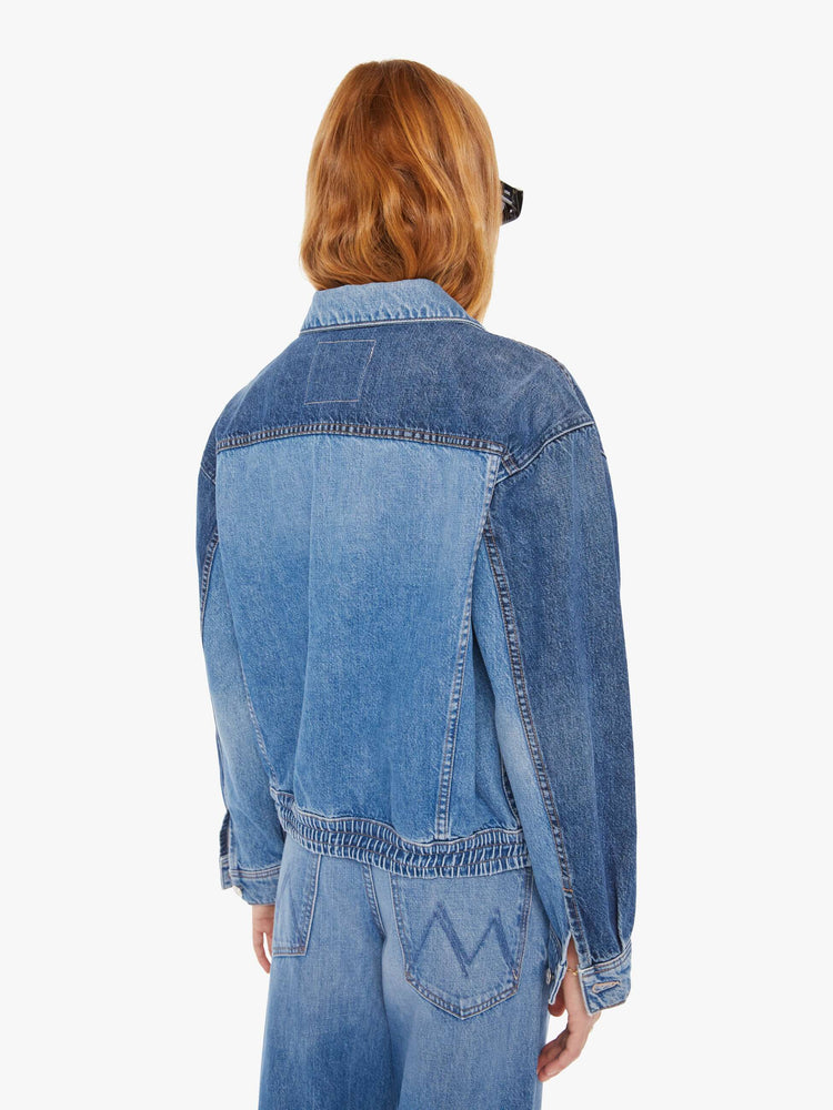 Back view of a woman mid blue denim jacket with drop shoulders, patch pockets and an elastic hem at the waist with darker details on chest.
