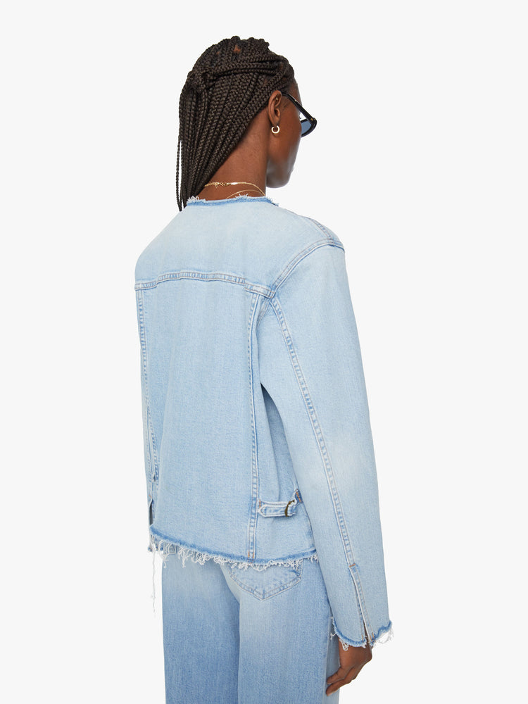 Back view of a women distressed denim jacket is designed with a crewneck, patch pockets, pleated details and raw, frayed hems in a light blue wash with missing patch pockets.