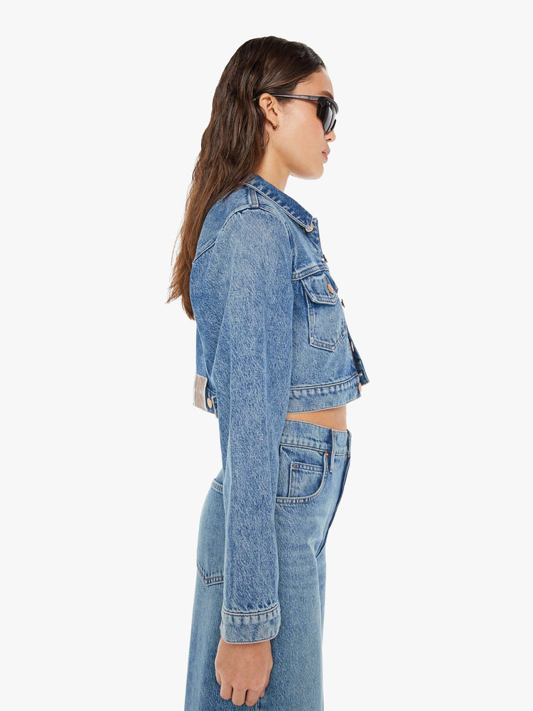 Side view of a woman wearing a medium blue wash denim jacket featuring a cropped fit, paired with a pair of wide leg jeans in the same color wash.