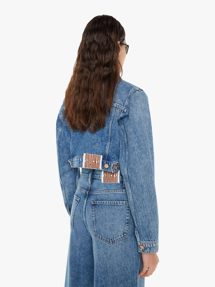 Back view of a woman wearing a medium blue wash denim jacket featuring a cropped fit, paired with a pair of wide leg jeans in the same color wash.