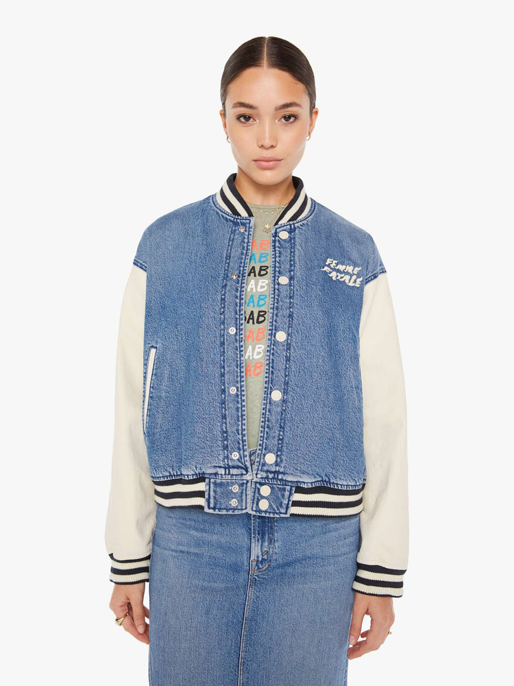 Front view of a womens denim varsity jacket featuring contrast with sleeves, black a white striped rib, white snap buttons, and white embroidery on the chest pocket reading "FEMME FATALE".