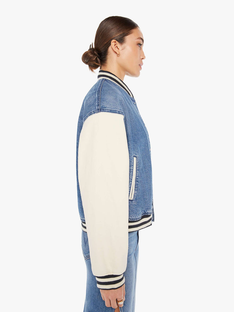 Side view of a womens denim varsity jacket featuring contrast with sleeves, black a white striped rib, white snap buttons, and white embroidery on the chest pocket reading "FEMME FATALE".