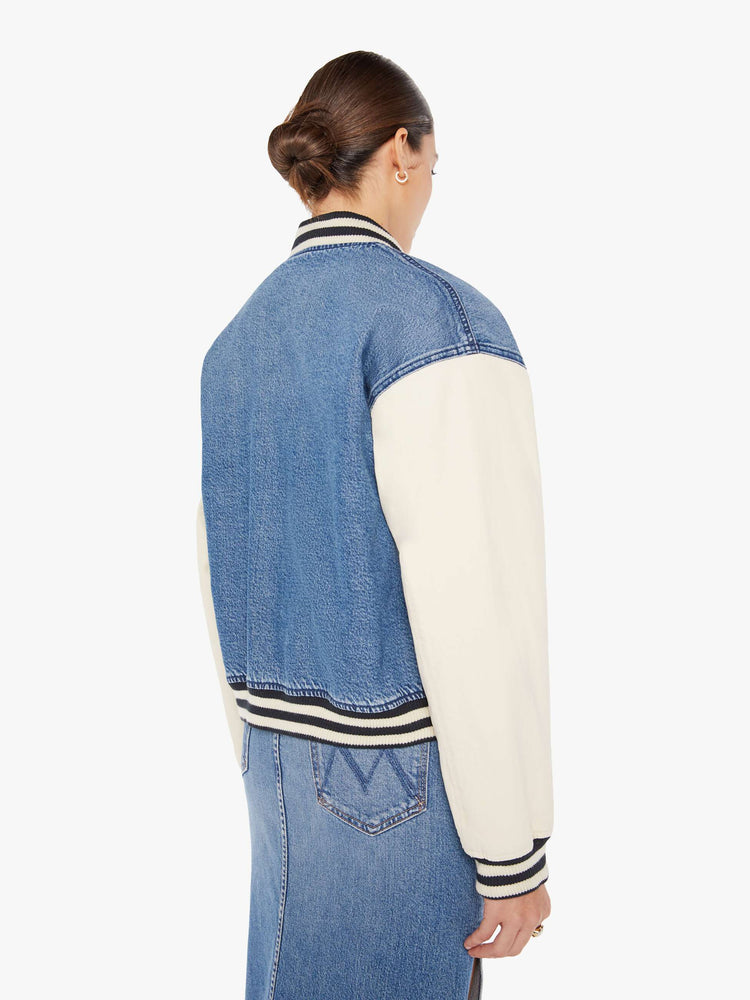 Back view of a womens denim varsity jacket featuring contrast with sleeves, black a white striped rib, white snap buttons, and white embroidery on the chest pocket reading "FEMME FATALE".