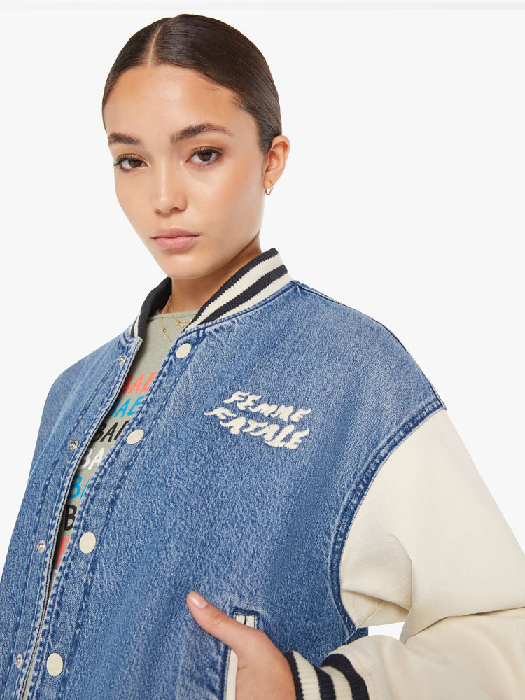 Front close up view of a womens denim varsity jacket featuring contrast with sleeves, black a white striped rib, white snap buttons, and white embroidery on the chest pocket reading "FEMME FATALE".