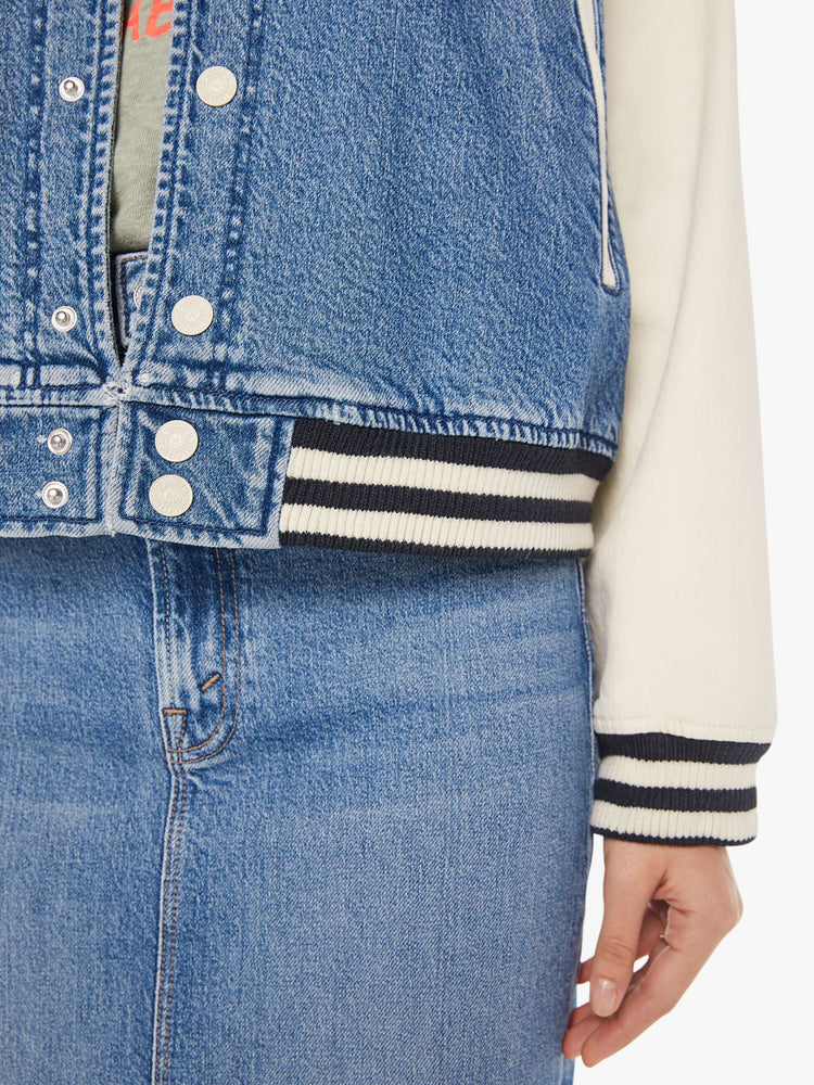 Front close up view of a womens denim varsity jacket featuring contrast with sleeves, black a white striped rib, and white snap buttons.