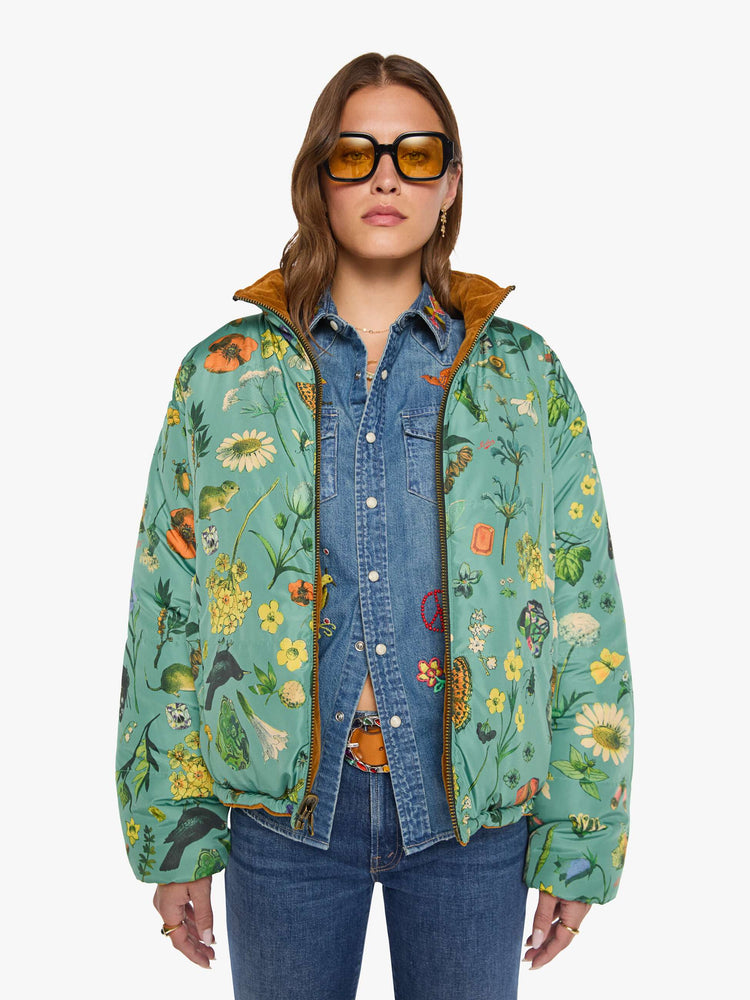 Front view of a woman in a  golden-brown corduroy on one side and a teal-green fabric on the other with oversized printed flowers, gems, and creatures throughout oversized puffer.