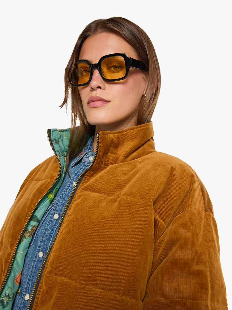 Close up view of a woman in a  golden-brown corduroy on one side and a teal-green fabric on the other with oversized printed flowers, gems, and creatures throughout oversized puffer.
