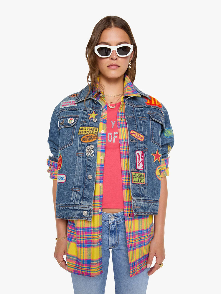 Front view of a woman in a mid blue with vintage patches oversized denim jacket with front patch pockets, drop shoulders, pleated details and a slightly cropped, boxy fit.
