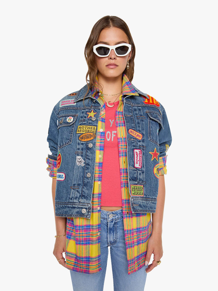 Front view of a woman in a mid blue with vintage patches oversized denim jacket with front patch pockets, drop shoulders, pleated details and a slightly cropped, boxy fit.
