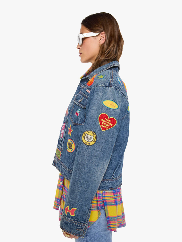 Left side view of a woman in a mid blue with vintage patches oversized denim jacket with front patch pockets, drop shoulders, pleated details and a slightly cropped, boxy fit.