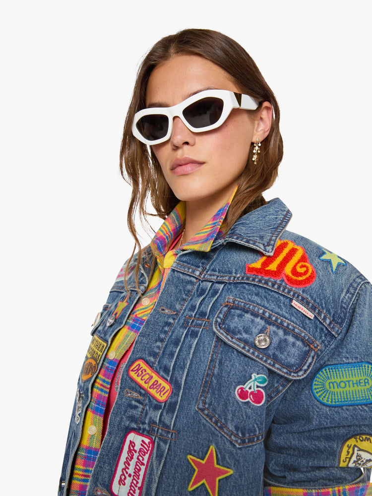 Close up view of a woman in a mid blue with vintage patches oversized denim jacket with front patch pockets, drop shoulders, pleated details and a slightly cropped, boxy fit.