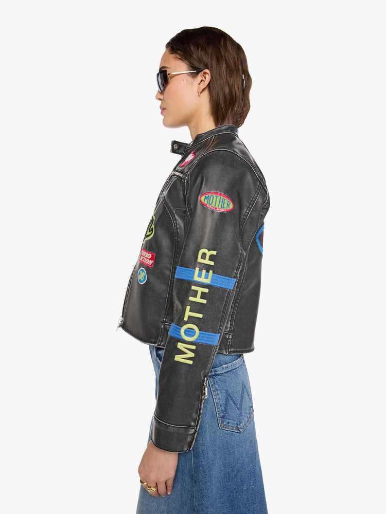 Side view of a woman in faux black leather with patches jacket with a button-tab crewneck, zip pockets, seamed details, a zip closure and a cropped fit.