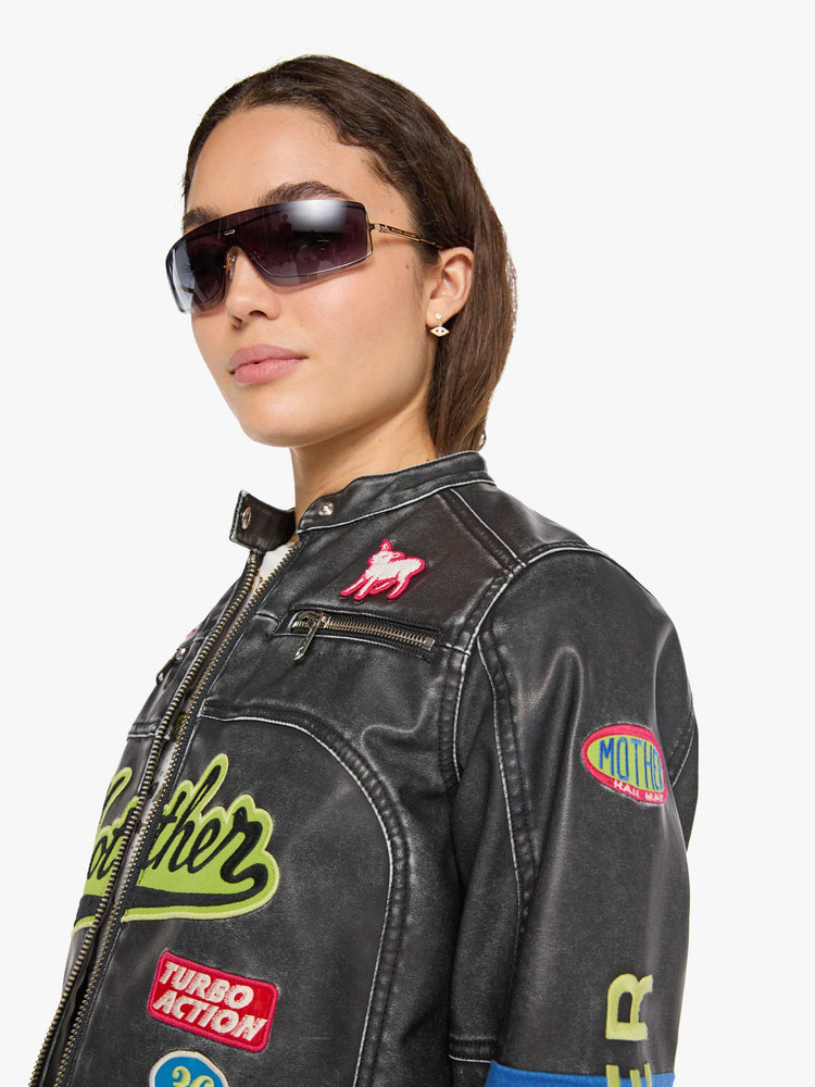 Close up view of a woman in faux black leather with patches jacket with a button-tab crewneck, zip pockets, seamed details, a zip closure and a cropped fit.
