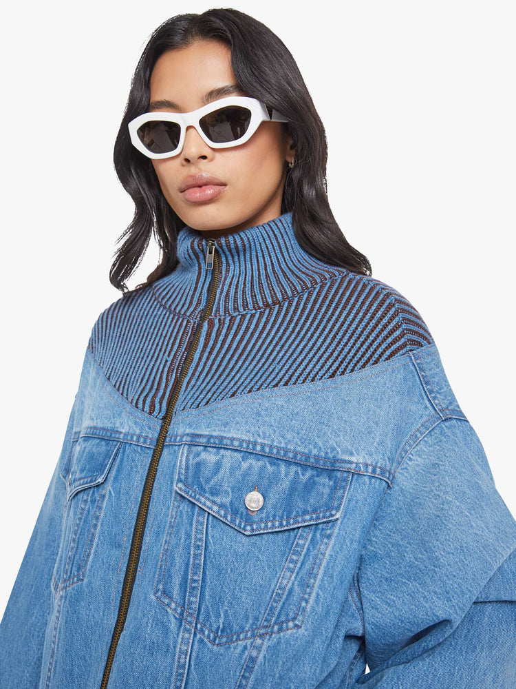 Close up view of a woman in a half denim jacket, half sweater with ribbed details around the neck, chest and shoulders, a high collar, drop shoulders, extra-wide long sleeves, front patch pockets and a super boxy fit. 