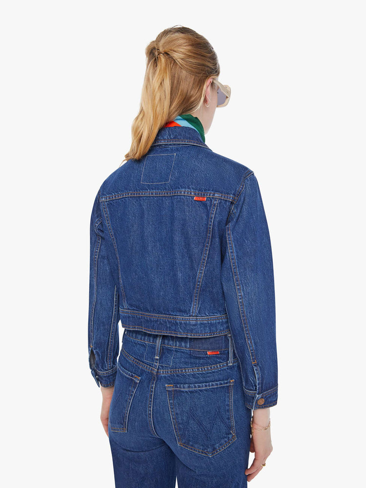Back view of a woman in a mid blue shrunken denim jacket with patch pockets and a cropped hem. 