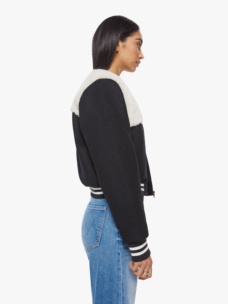 Side view of a woman dark blue with white faux-sherpa details on the shoulders and striped hem zip-up varsity bomber jacket.