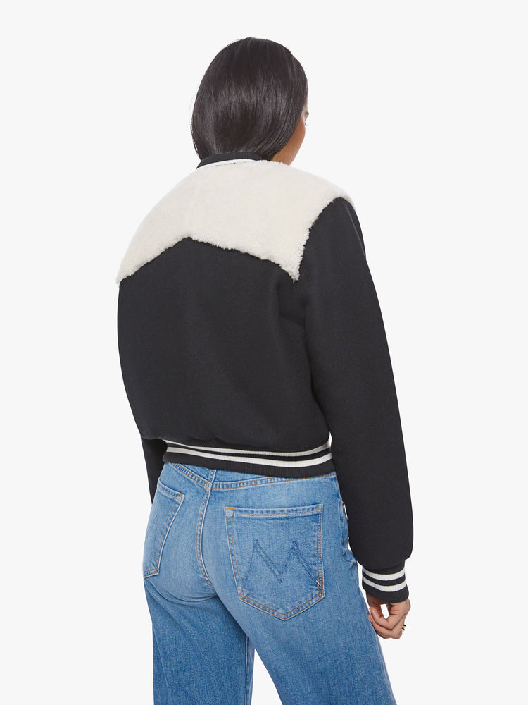 Back view of a woman dark blue with white faux-sherpa details on the shoulders and striped hem zip-up varsity bomber jacket.