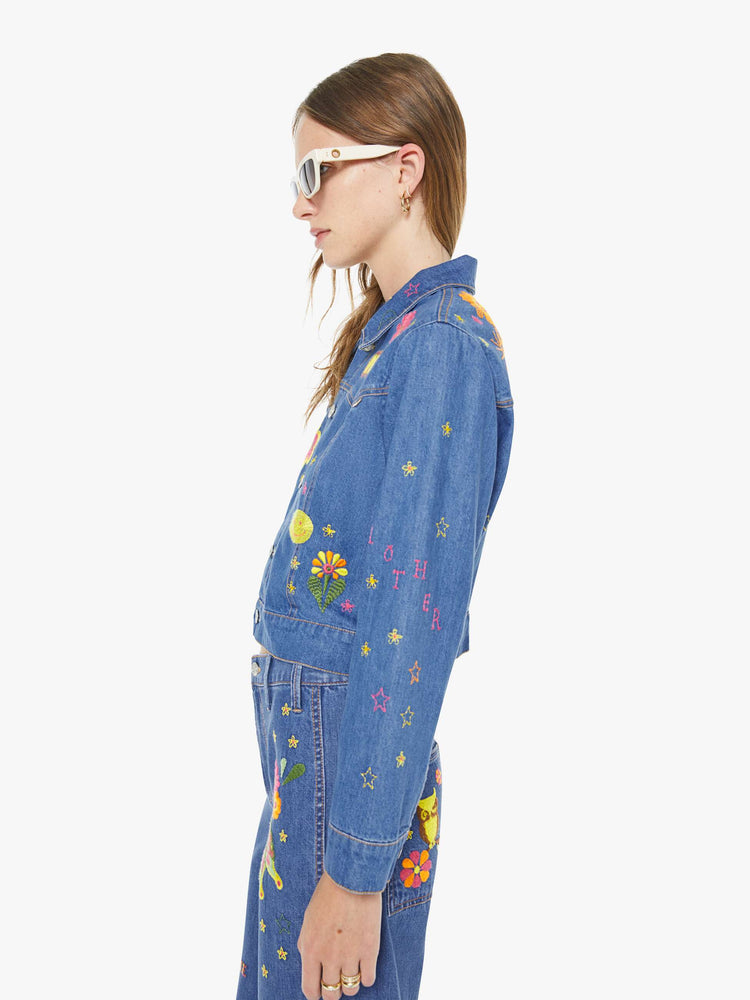 Left side view of a woman in a medium blue with embroidery flowers denim jacket with seamed details and a cropped fit.