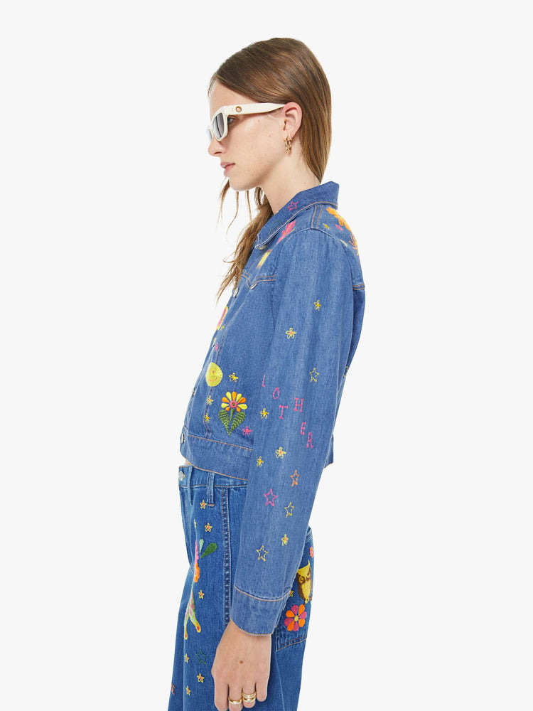 Left side view of a woman in a medium blue with embroidery flowers denim jacket with seamed details and a cropped fit.