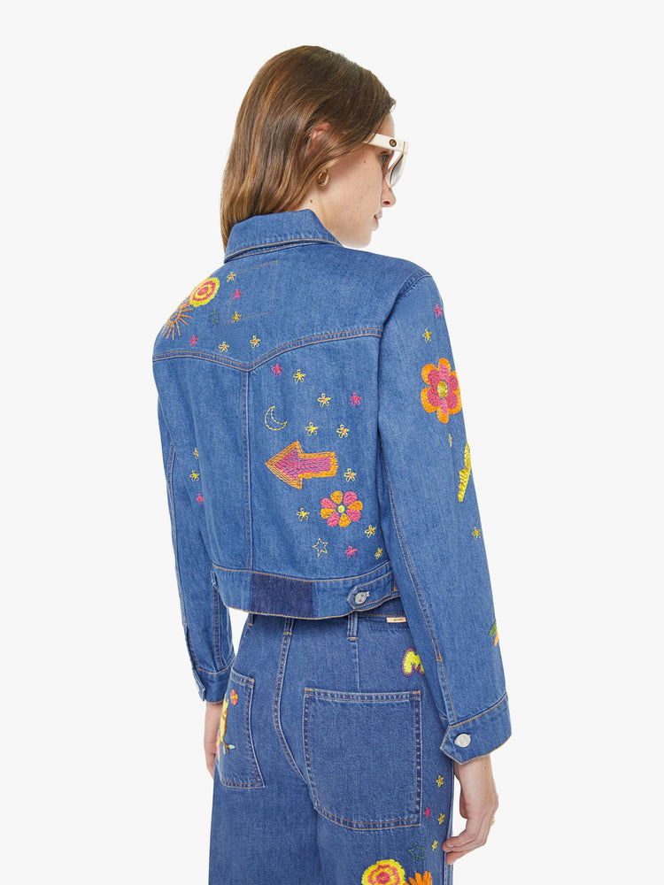 Back view of a woman in a medium blue with embroidery flowers denim jacket with seamed details and a cropped fit.
