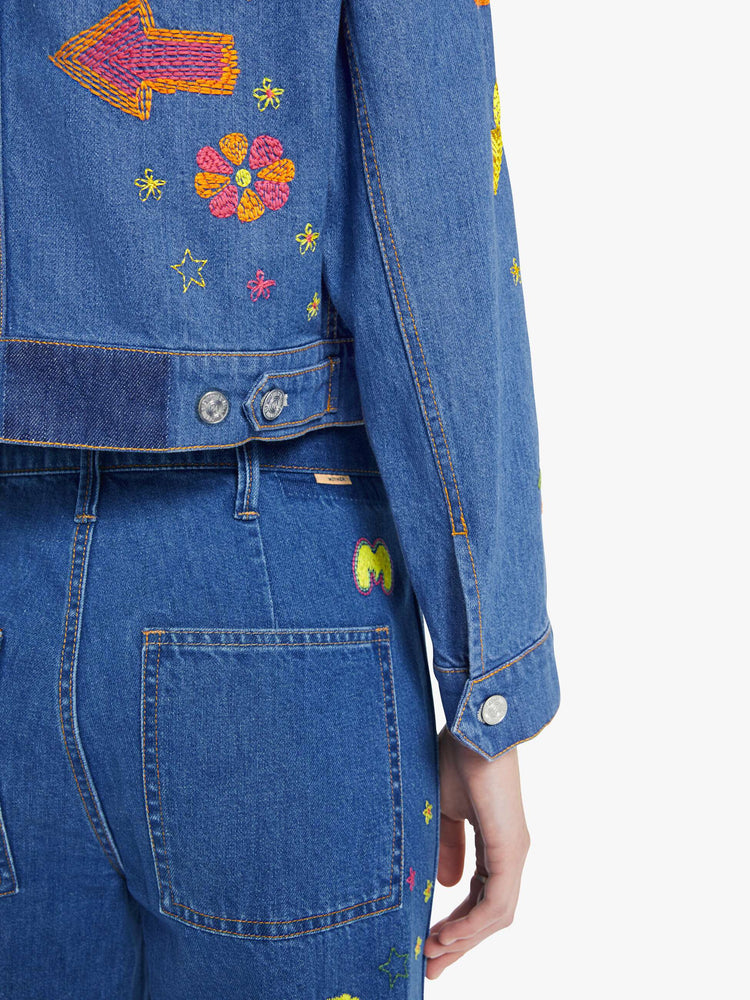 Swatch view of a woman in a medium blue with embroidery flowers denim jacket with seamed details and a cropped fit.