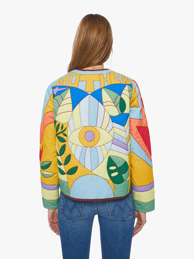 Back view of a woman quilted jacket with a crewneck, curved hems, hook closures down the front and a boxy fit in a colorful quilted fabric reminiscent of stained glass.