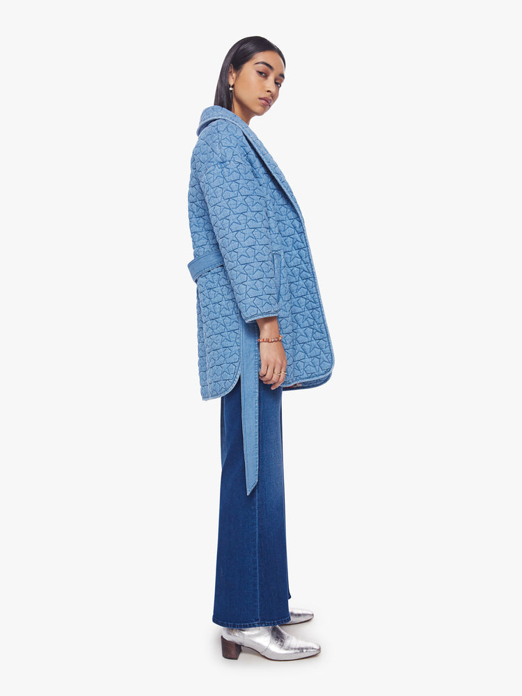 Side view of a woman quilted jacket with a shawl collar, drop shoulders, belted waist, side slit pockets and a thigh-grazing hem in a soft blue hue stars and colorful floral lining.