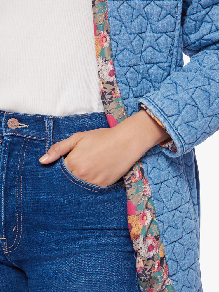Close up view of a woman quilted jacket with a shawl collar, drop shoulders, belted waist, side slit pockets and a thigh-grazing hem in a soft blue hue stars and colorful floral lining.