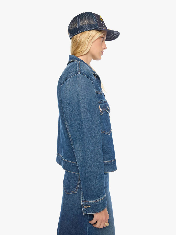 Side view of a woman in a med blue denim jacket with a Western-inspired yoke, sawtooth pointed patch pockets on the chest and a cropped, boxy fit.