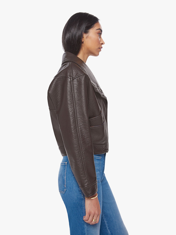 Side view of a woman brown faux leather jacket with a dramatically oversized collar, front slit pockets, drop shoulders and a hidden zip closure with buttons down the front.