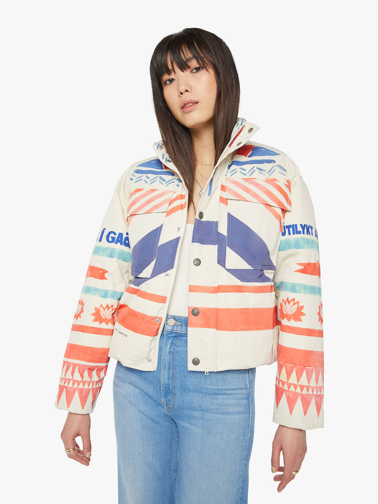 Front view of a woman weatherproof puffer jacket with a high neck, oversized patch pockets, side slit pockets, a hidden zip closure that buttons and double-layer hems in creamy with symbolic graphics and text in red and blue.