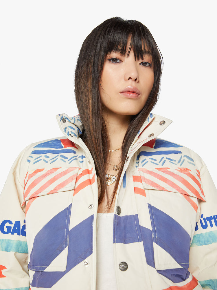 Close up view of a woman weatherproof puffer jacket with a high neck, oversized patch pockets, side slit pockets, a hidden zip closure that buttons and double-layer hems in creamy with symbolic graphics and text in red and blue.